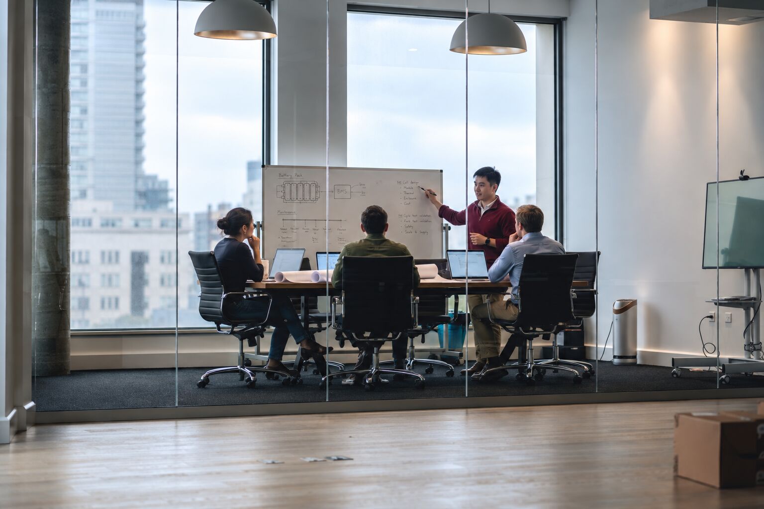 Four engineers in a glass-walled meeting room. One points to a sketch of a battery pack and its BMS on a whiteboard.
